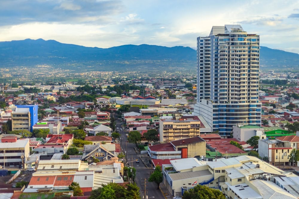 A cityscape with a tall modern building surrounded by smaller structures, tree-lined streets, and mountains in the…