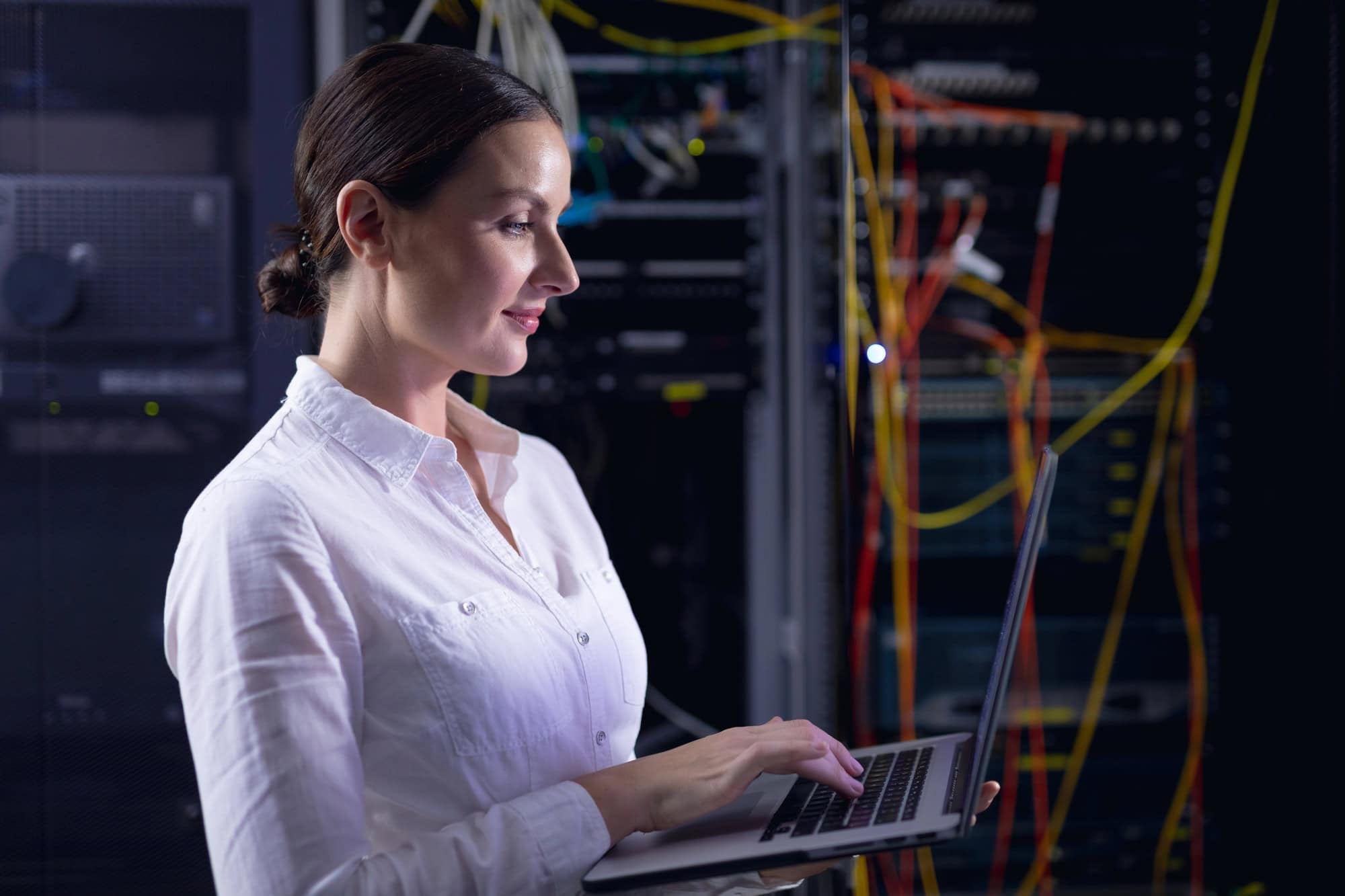 A woman in a white shirt is using a laptop while standing in a server room, managing custom cloud infrastructure with IT…