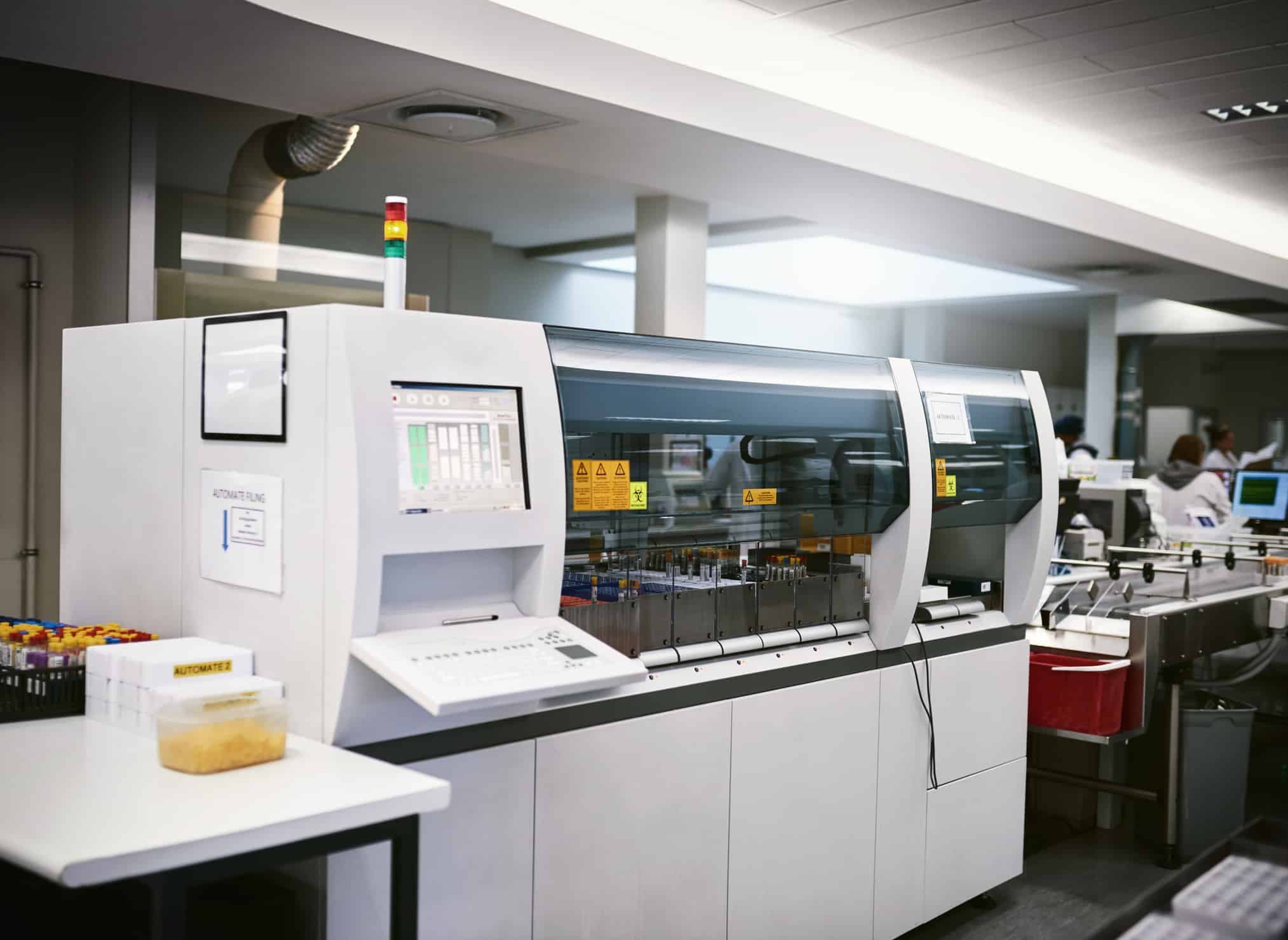 A large automated laboratory machine processes samples in a secure biotech lab, with tubes and containers visible nearby…